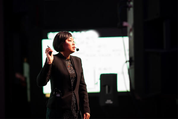Master Suyen Lim presenting on a darkened stage with a headset microphone